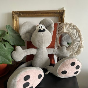 Könnte beinhalten: Ein graues Plüschmaus-Spielzeug mit großen Ohren, einer schwarzen Nase und rosa und schwarzen Füßen. Das Spielzeug liegt auf einer schwarzen Oberfläche. Ein goldgerahmtes Bild und ein dekorativer Wandschmuck befinden sich im Hintergrund.