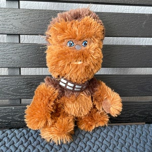 Op de afbeelding: Een bruin, pluche Chewbacca-speelgoed met blauwe ogen en een bruine bandoleer met witte strepen. Het speelgoed zit op een blauw, getextureerd oppervlak.