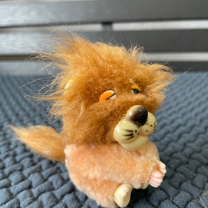 Puede incluir: Un pequeño juguete de peluche de león con pelo marrón y nariz rosa. El león está sentado con las patas juntas y tiene un ojo cerrado.