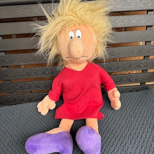 May include: A plush doll with a light skin tone, blonde hair, and a red dress. The doll has large purple shoes and is sitting on a dark gray surface. The doll's facial features include a large nose and wide eyes.