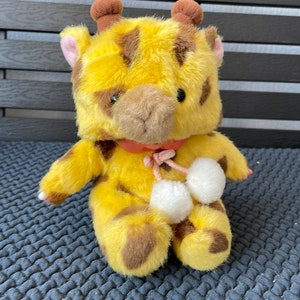 May include: A yellow and brown plush giraffe toy with white pom-poms on its feet. The giraffe has a pink bow around its neck.
