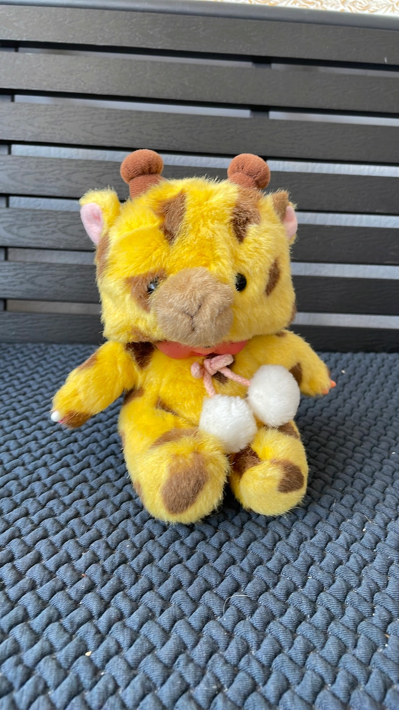May include: A yellow and brown plush giraffe toy with white pom-poms on its feet. The giraffe has a pink bow around its neck.