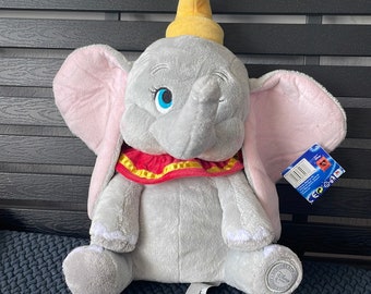 May include: A gray plush Dumbo the elephant toy with a yellow hat, red and yellow collar, and blue eyes. The toy is sitting on a dark gray textured surface.