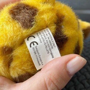 May include: A yellow and brown plush giraffe toy with a white fur tail. The toy has a label that reads "New Materials Only Reg. No. PA-10005(CN) Contents: Polyester Fibers and Plastic PE Pellets Made in China CE Sekiguchi Europe b.v. 3621BK-111/Holland".