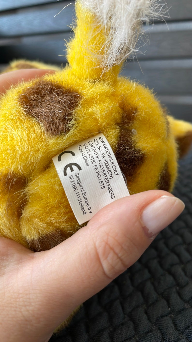 May include: A yellow and brown plush giraffe toy with a white fur tail. The toy has a label that reads "New Materials Only Reg. No. PA-10005(CN) Contents: Polyester Fibers and Plastic PE Pellets Made in China CE Sekiguchi Europe b.v. 3621BK-111/Holland".