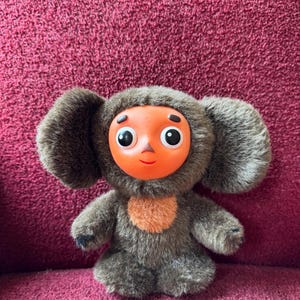 Op de afbeelding: Een pluche speelgoed met een bruin, harig lichaam, grote ronde oren en een fel oranje gezicht met grote zwarte ogen. Het speelgoed heeft een kleine oranje vlek op de borst en staat op een rode ondergrond.