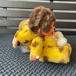 May include: A yellow and brown plush monkey toy wearing a yellow and brown spotted outfit. The monkey is sitting on a gray textured surface.