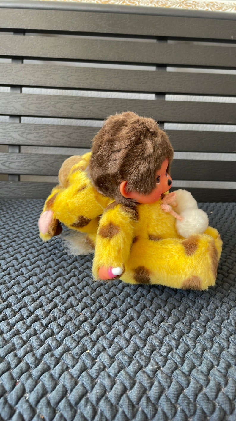 May include: A yellow and brown plush monkey toy wearing a yellow and brown spotted outfit. The monkey is sitting on a gray textured surface.