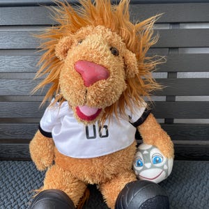 Könnte beinhalten: Ein Plüschlöwe, der ein weißes Fußballtrikot mit der Nummer 08 trägt. Der Löwe hält einen Fußball mit einem lächelnden Gesicht. Der Löwe sitzt auf einer grauen Oberfläche.