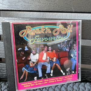 Puede incluir: Una caja de CD con fondo rosa y el texto "Rock 'n' Roll Favourites" en una fuente colorida. La imagen muestra a un grupo de personas sentadas en un cine, disfrutando de una película. El texto en la parte inferior de la caja enumera las canciones incluidas en el CD: "Hound Dog", "Tutti Frutti", "Rock Around The Clock", "Yakety Yak", "Rubber Ball", "Be Bop A Lula", "Johnny B. Goode", y "many others".