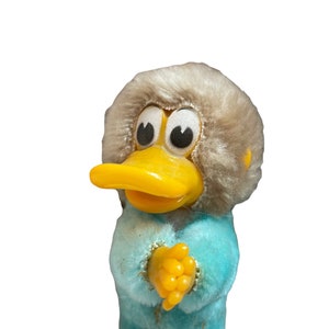 Puede incluir: Un pato de peluche que lleva un mono azul y un gorro de piel gris. El pato tiene grandes ojos amarillos y un pico amarillo.