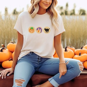 May include: A white t-shirt with three orange, yellow, and purple circles featuring a pumpkin, ghost, and black cat graphic. The shirt is worn by a person wearing blue jeans.