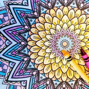Könnte beinhalten: Ein detailliertes Mandala-Design mit einer gelben Blume in der Mitte. Das Mandala ist in Blautönen, Rosa und Lila gefärbt. Zwei Kreiden, eine gelbe und eine rosa, liegen auf dem Mandala.