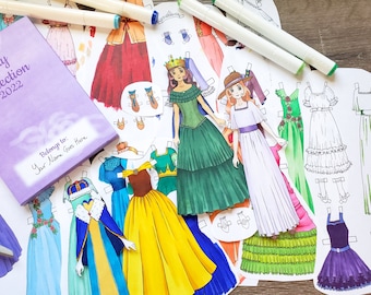 Peut inclure: Un livret violet avec le texte "Filly Collection 2022" et "Appartient à : Votre nom ici". Le livret est entouré de poupées en papier avec différentes robes et accessoires, dont une robe verte avec une couronne, une robe blanche avec un chapeau et une robe violette avec une ceinture noire.