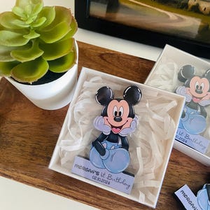 Puede incluir: Figura de Mickey Mouse en una caja blanca con el texto "Morgan's 1st Birthday 02.10.2024". La figura es negra, blanca y azul. La caja está sobre una bandeja de madera con una pequeña planta verde.