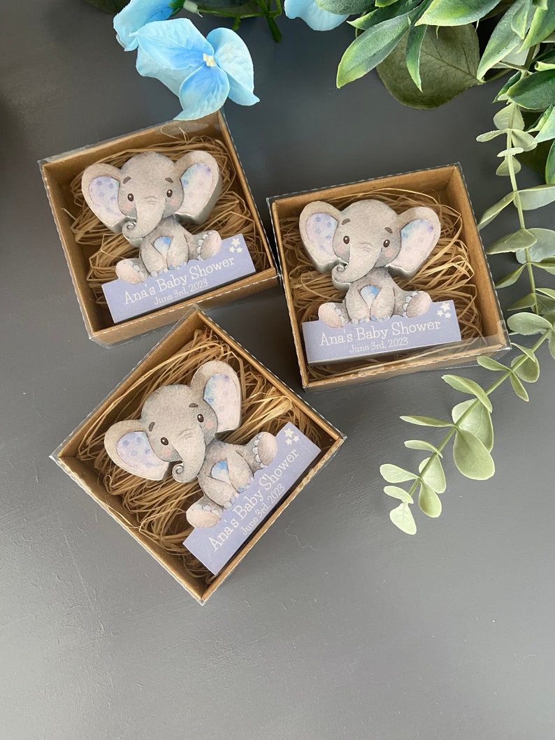 Op de afbeelding: Drie kleine, transparante doosjes met bruine deksels gevuld met gescheurd papier. Elk doosje bevat een klein, grijs olifantbeeldje met blauwe accenten. De doosjes hebben een paars label met de tekst "Ana's Baby Shower June 3rd, 2023".