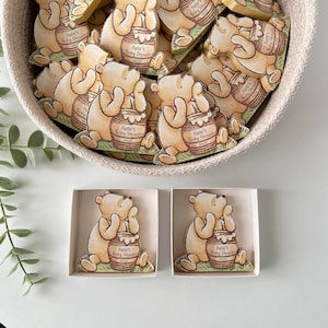 May include: A set of wooden Winnie the Pooh-themed baby shower favors. Each favor features a cartoon Winnie the Pooh holding a honey pot with the text "Hallie's Baby Shower" on it. The favors are in a basket and two are in individual boxes.