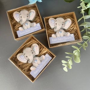 Op de afbeelding: Drie kleine, transparante doosjes met een lichtbruine basis bevatten een houten olifantenfiguur met blauwe en witte accenten. Elk doosje heeft een klein bordje met de tekst "Ana's Baby Shower" en de datum "June 3rd, 2023".