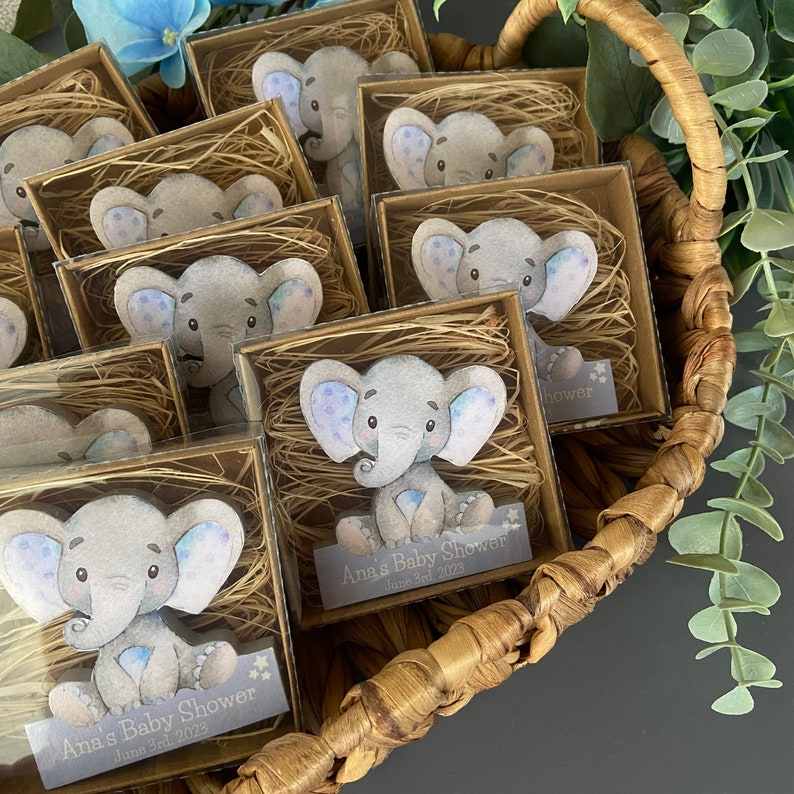 Op de afbeelding: Een mand gevuld met kleine doosjes die een grijs olifantbeeldje met blauwe accenten bevatten. Elk doosje heeft een label met de tekst "Ana's Baby Shower" en de datum "June 3rd, 2023".
