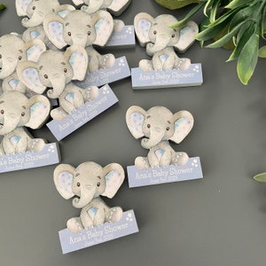Op de afbeelding: Een set blauwe en grijze houten olifant baby shower-cadeautjes met een wit sterrenontwerp. Elke olifant houdt een bord vast met de tekst "Ana's Baby Shower" en de datum "June 3rd, 2023".