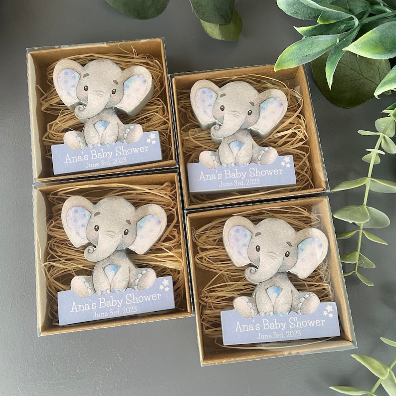 Op de afbeelding: Vier kleine cadeaudoosjes met doorzichtige deksels, elk met een handgeschilderd houten olifantbeeldje. De doosjes zijn gevuld met gescheurd papier en elk heeft een blauw en wit label met de tekst "Ana's Baby Shower June 3rd, 2023".