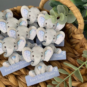 Op de afbeelding: Een mand gevuld met blauwe en grijze olifant baby shower-cadeautjes. Elke olifant heeft blauwe en witte stippen op zijn oren. De cadeautjes hebben een blauw en wit bord met de tekst "Ana's Baby Shower June 3rd, 2023".