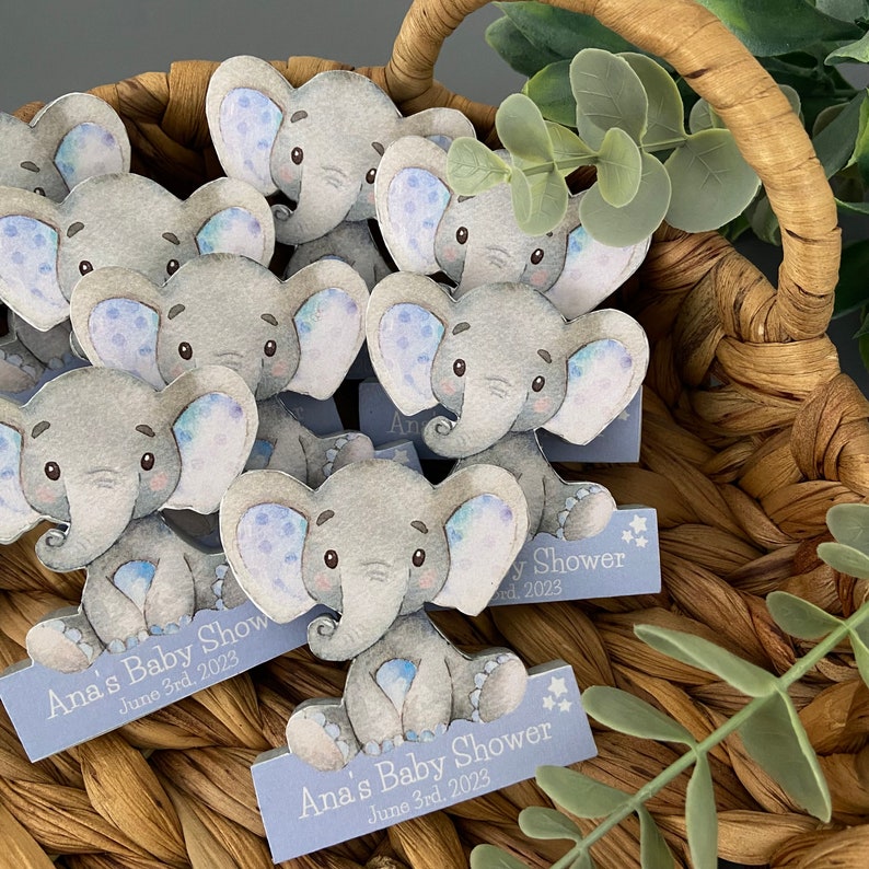 Op de afbeelding: Een mand gevuld met blauwe en grijze olifant baby shower-cadeautjes. Elke olifant heeft blauwe en witte stippen op zijn oren. De cadeautjes hebben een blauw en wit bord met de tekst "Ana's Baby Shower June 3rd, 2023".