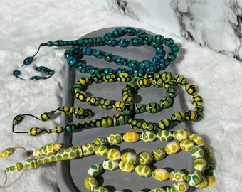 May include: Three strands of prayer beads featuring oval-shaped beads with a distinct honeycomb pattern. The top strand is blue and black, the middle is yellow and green, and the bottom is white, yellow, and green. Each strand ends with a tassel.