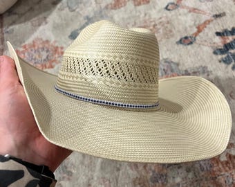 Könnte beinhalten: Ein hellbeiger Strohhut im Cowboy-Stil mit einem blauen und weißen Band um die Krone.