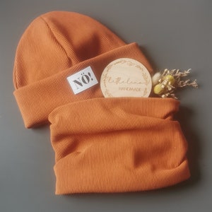 Könnte beinhalten: Ein Set aus zwei orangefarbenen gerippten Strickmützen, eine Mütze und ein Halstuch. Die Mütze hat ein weißes Etikett mit dem Wort "NÖ!" darauf. Der Halstuch hat ein Holzschild mit dem Text "littlelino HANDMADE" darauf.