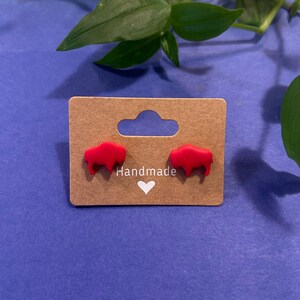 May include: A pair of red buffalo-shaped stud earrings displayed on a brown card with the word "Handmade" and a white heart. The earrings are set against a blue background with green foliage.