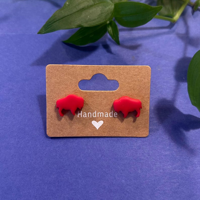 May include: A pair of red buffalo-shaped stud earrings displayed on a brown card with the word "Handmade" and a white heart. The earrings are set against a blue background with green foliage.