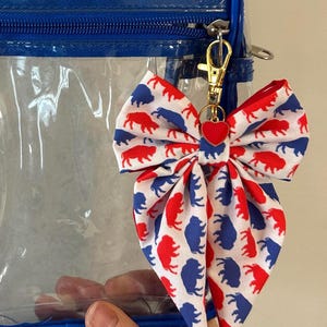 May include: A red, white, and blue bow-shaped keychain with a heart charm, attached to a clear blue-trimmed bag. The bow features a repeating pattern of red and blue bison on a white background. The keychain is attached with a gold-colored clasp.
