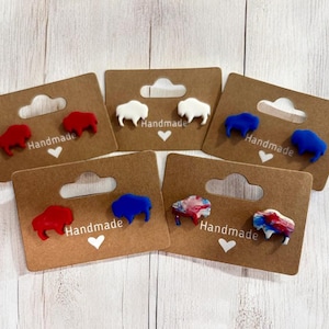 May include: Handmade buffalo-shaped stud earrings in red, white, blue, and multi-color designs. Each pair is mounted on a brown card with a heart and the word "Handmade".