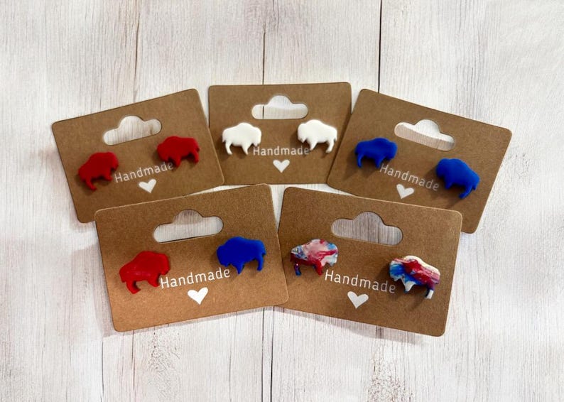 May include: Handmade buffalo-shaped stud earrings in red, white, blue, and multi-color designs. Each pair is mounted on a brown card with a heart and the word "Handmade".