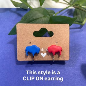 May include: Clip-on earrings featuring a blue and a red buffalo design. The earrings are displayed on a brown card with the text "This style is a CLIP ON earring" on a blue background with green leaves.