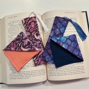 May include: Two fabric bookmarks with a corner pocket. One bookmark features a purple and blue watercolor dragon scale pattern with a navy blue corner pocket. The other bookmark features a pink and purple watercolor dragon pattern with a coral corner pocket.