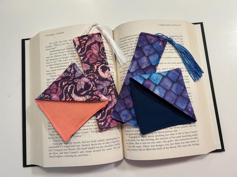 May include: Two fabric bookmarks with a corner pocket. One bookmark features a purple and blue watercolor dragon scale pattern with a navy blue corner pocket. The other bookmark features a pink and purple watercolor dragon pattern with a coral corner pocket.