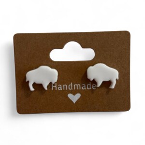May include: White buffalo-shaped earrings on a brown card with the word "Handmade" and a heart. The earrings are small and have a smooth finish. The card has a hole at the top for hanging.