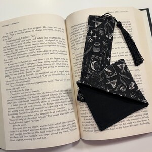 May include: A black and white bookmark with a tassel. The bookmark features a repeating pattern of moons, stars, and other celestial imagery. The bookmark is lying on top of an open book.