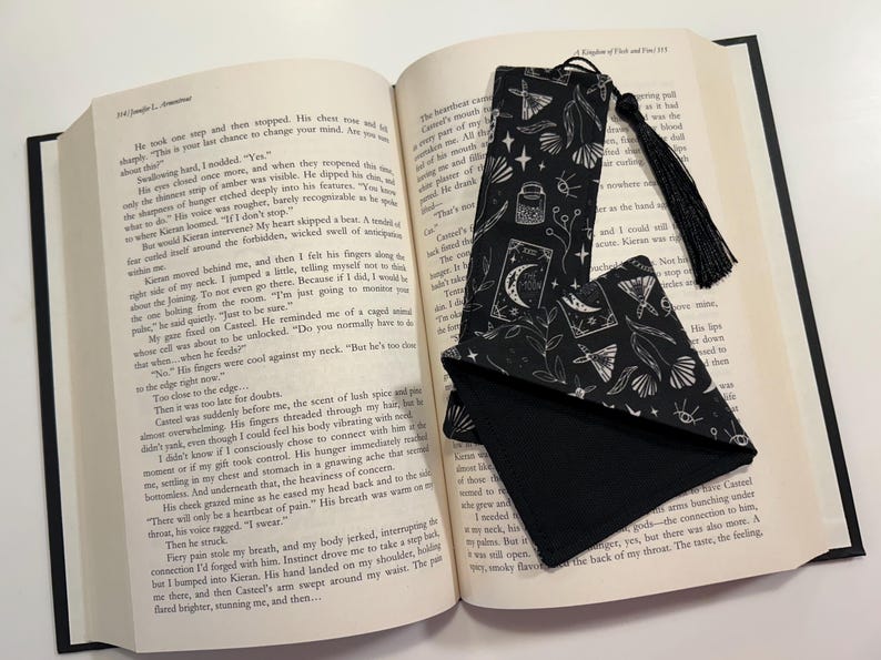 May include: A black and white bookmark with a tassel. The bookmark features a repeating pattern of moons, stars, and other celestial imagery. The bookmark is lying on top of an open book.
