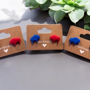 May include: Three pairs of handmade stud earrings featuring buffalo shapes in red and blue. Each pair is mounted on a brown card with the word "Handmade" and a heart symbol. The earrings are displayed on a gray surface.