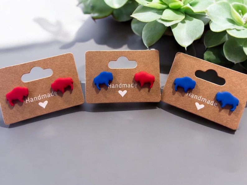 May include: Three pairs of handmade stud earrings featuring buffalo shapes in red and blue. Each pair is mounted on a brown card with the word "Handmade" and a heart symbol. The earrings are displayed on a gray surface.