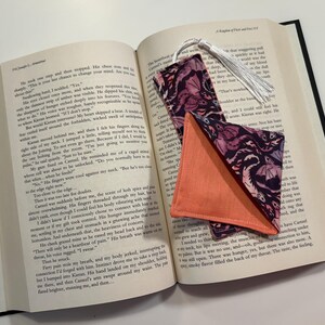 May include: Two fabric bookmarks with a pink and black floral pattern. One bookmark is a rectangle with a tassel and the other is a triangle. Both bookmarks are folded and placed on an open book.