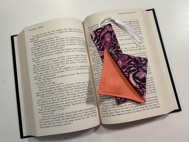 May include: Two fabric bookmarks with a pink and black floral pattern. One bookmark is a rectangle with a tassel and the other is a triangle. Both bookmarks are folded and placed on an open book.