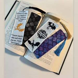 May include: Four fabric bookmarks with dragon designs and quotes. The bookmarks are on a book with a blue tassel hanging off the bottom of one bookmark. The quotes on the bookmarks are "A dragon without its rider is a tragedy. A rider without their dragon is dead." and "I know exactly who and what you are, Violet Sorrengail."