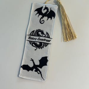 May include: White fabric bookmark with a gold tassel. The bookmark features three black dragon silhouettes and the text "Riders Quadrant".