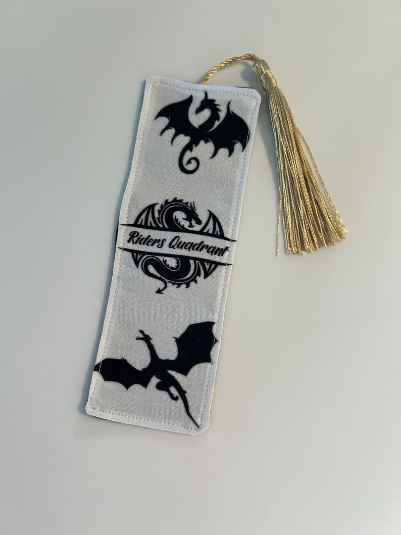 May include: White fabric bookmark with a gold tassel. The bookmark features three black dragon silhouettes and the text "Riders Quadrant".