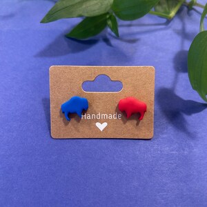 May include: A pair of handmade stud earrings featuring buffalo shapes, one blue and one red. The earrings are displayed on a brown card with the word "Handmade" and a heart symbol. The background is a solid blue.