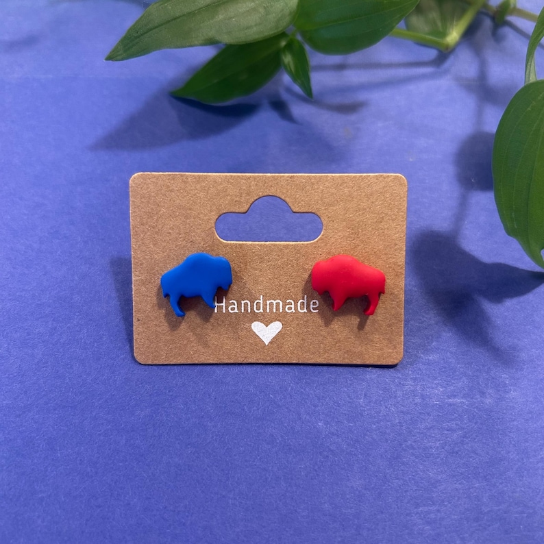 May include: A pair of handmade stud earrings featuring buffalo shapes, one blue and one red. The earrings are displayed on a brown card with the word "Handmade" and a heart symbol. The background is a solid blue.